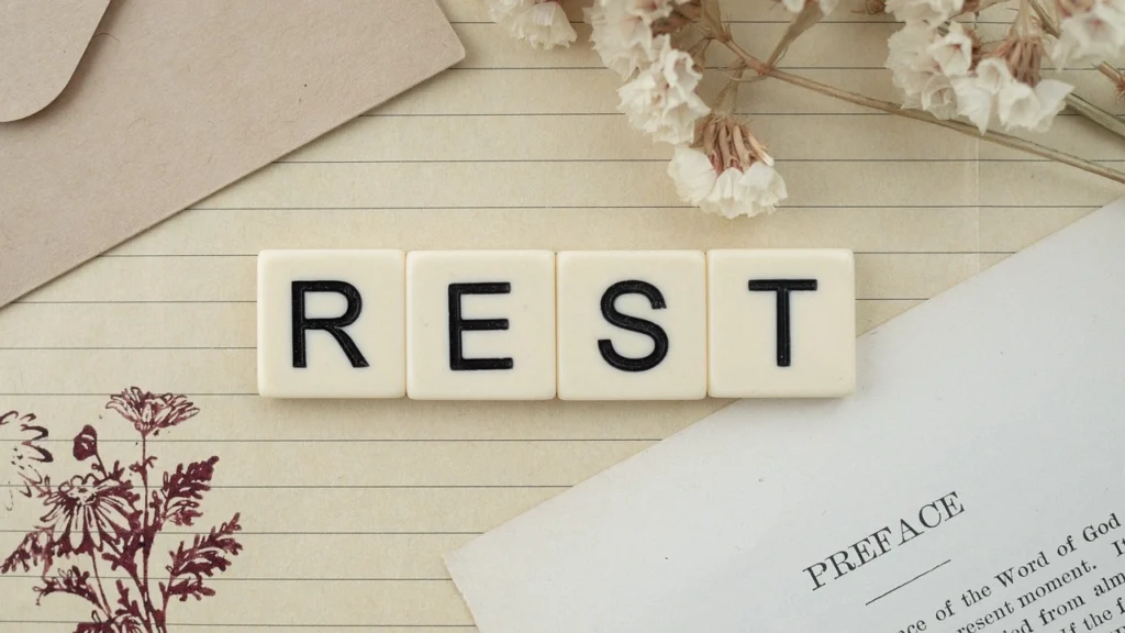 המילה "REST" כתובה באותיות בולטות על רקע עיצוב טבעי ועדין, עם פרחים יבשים וניירות כתיבה המשרים תחושת רוגע ושלווה. התמונה מדגישה את החשיבות של מנוחה איכותית עבור הבריאות הנפשית והפיזית.